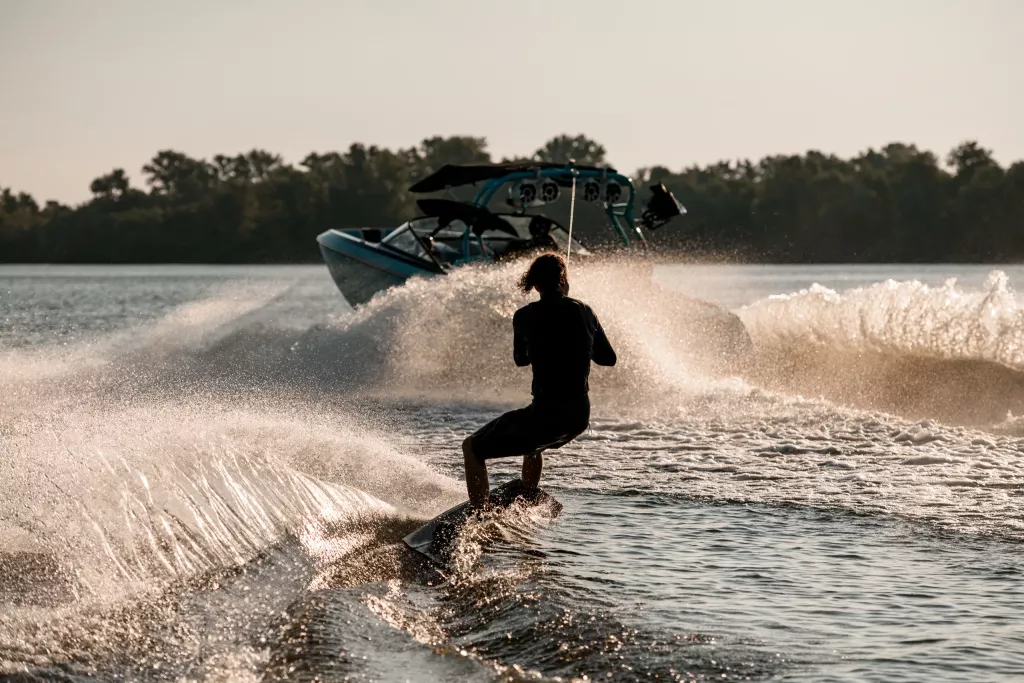 Rear view of athletic guy holding rope and energetically riding wakeboard behind motor boat on splashing river waves. Active and extreme sports