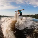 Young energy male wakesurf rider jumping on the waves of the river against the blue sky and trees