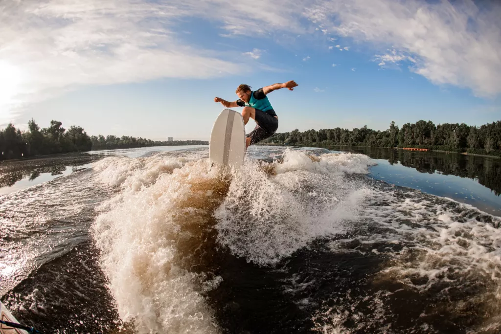 Young energy male wakesurf rider jumping on the waves of the river against the blue sky and trees