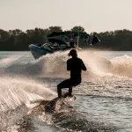Rear view of athletic guy holding rope and energetically riding wakeboard behind motor boat on splashing river waves. Active and extreme sports